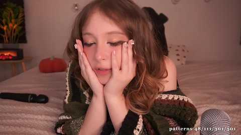 Media: Video of a young Caucasian girl with fair skin and long brown hair, lying on a bed with a white blanket. She wears a green knitted sweater and is playfully sticking out her tongue.