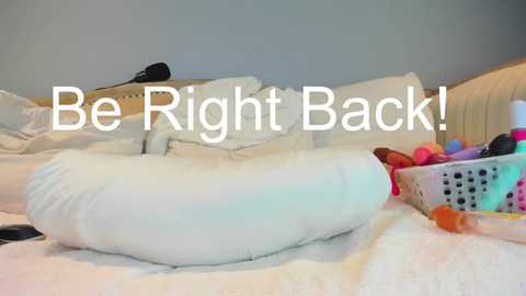 Media: Video of a messy bedroom with white bedding, a pillow, a toy basket, and a black hairbrush on the bed, overlaid with white text reading \"Be Right Back!\