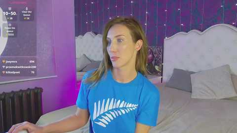 Media: Video of a Caucasian woman with light skin and shoulder-length brown hair, wearing a blue t-shirt, standing in a modern bedroom with a white tufted headboard and purple-tinted curtains.