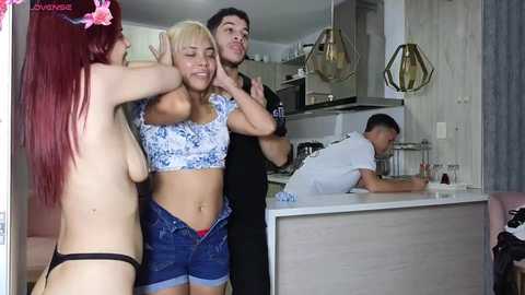 Media: Video of a red-haired woman topless, wearing only black thong, standing next to a blonde woman in a floral crop top and denim shorts. A shirtless man in black stands behind them, and another man in white leans over a kitchen counter.