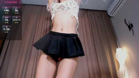 Media: Video of a slender, light-skinned woman in a short, black skirt, revealing her black panties and flat stomach. She's indoors with beige curtains, a white wall, and a ceiling fan.