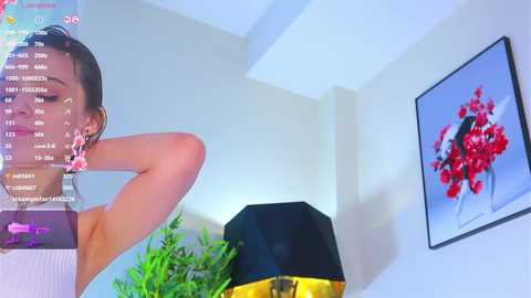 Media: Video of a woman with brown hair, wearing a white top, sitting in a room with a black lamp, green plant, and a red flower painting on the wall.
