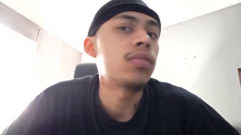 Media: A video of a young Latino man with medium brown skin, wearing a black baseball cap, black T-shirt, and serious expression, sitting in a dimly lit room with white walls.