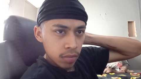 Media: A video of a young Black man with a light brown complexion, wearing a black cap and T-shirt, sitting in a black chair, with a casual, neutral expression.