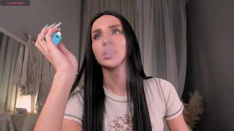 Video of a slender, fair-skinned woman with long black hair blowing a bubble. She's wearing a light gray T-shirt and holding a blue object. Background features draped curtains and a boho-style lamp.
