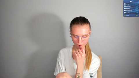 Media: Video of a blonde woman with glasses and a ponytail, wearing a white shirt, seated against a plain gray wall, appearing to be in a thoughtful or contemplative state.