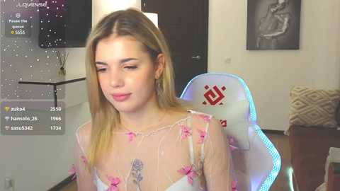 Media: Video of a young, fair-skinned blonde woman with long hair, wearing a sheer white dress with pink floral embroidery, sitting in a gaming chair in a modern, minimalist room with a black TV and abstract artwork on the wall.