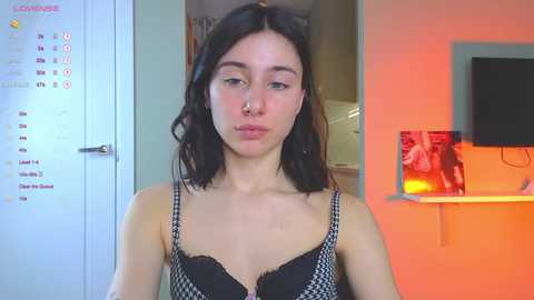 Media: A video of a young Caucasian woman with shoulder-length black hair, wearing a black and white patterned bra, standing indoors with a calendar, wall art, and a TV in the background.
