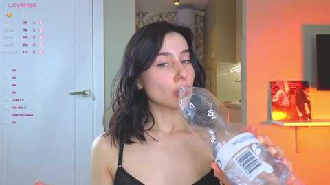 Media: A video of a young woman with fair skin, dark hair, and a slender physique, drinking from a clear plastic water bottle, in a brightly lit, modern kitchen.