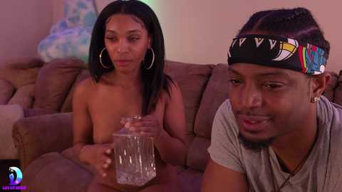 Media: A video of two young Black people on a couch, a woman with straight black hair, topless, holding a glass, and a man in a gray t-shirt with a black headband, both looking at the glass.