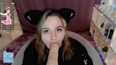 Media: Video of a young woman with long, wavy blonde hair, wearing black cat ears, kneeling on a plush chair, performing oral sex on a large erect penis.