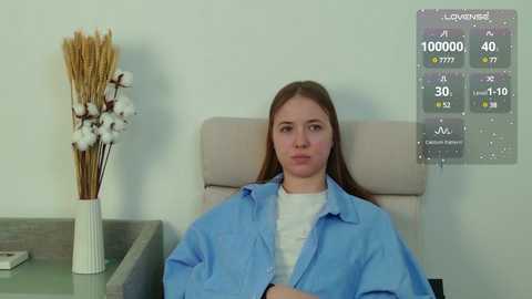 Video of a young woman with light skin and straight brown hair, wearing a blue surgical gown, seated in a beige chair. A white vase with dried flowers and a digital screen showing medical information are in the background.