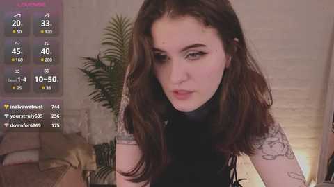 Media: Video of a young woman with long, dark brown hair, pale skin, and tattoos, wearing a black turtleneck, indoors with a green plant and a TV screen showing TikTok stats in the background.