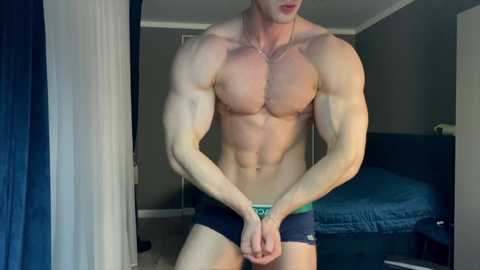 Media: Video of a muscular, shirtless man wearing dark blue briefs, standing indoors with gray walls, a blue bed, and a white door.