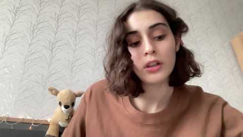 Media: Video of a young woman with wavy dark hair, wearing a brown shirt, standing in a room with a light wallpaper featuring tree patterns. She looks contemplative, with a stuffed animal and string lights in the background.