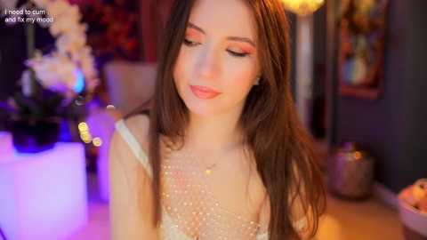 Media: A video of a young woman with long brown hair, wearing a sparkling white dress, smiling softly in a dimly lit room with blurred floral decorations and warm lighting.