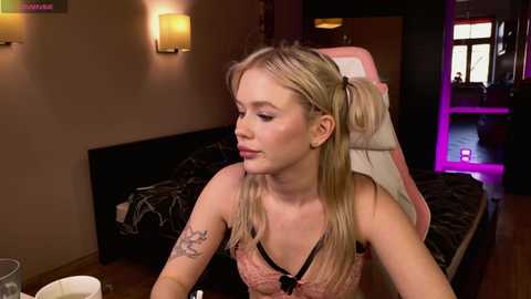 Video of a blonde woman with long hair in pigtails, wearing a pink lace top, sitting in a dimly-lit room with neon lights, looking at a phone.