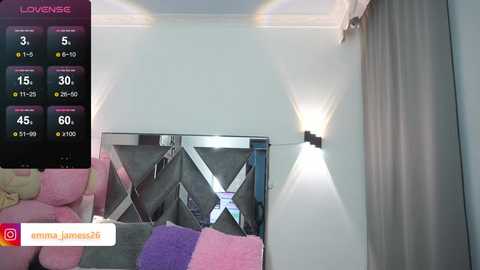 Media: Video of a modern living room with a large flat-screen TV, pink and purple throw pillows on a beige sofa, and a white wall-mounted light fixture.