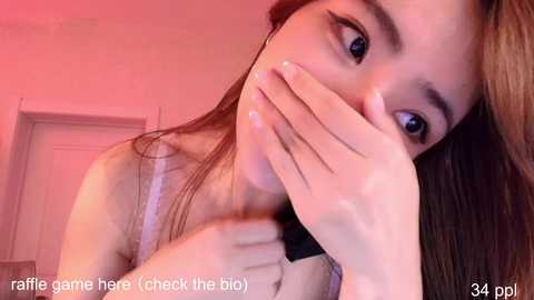 Media: A close-up video of a young Asian woman with fair skin, long brown hair, and a slender physique, wearing a white spaghetti-strap top. She covers her mouth with her hand, looking surprised.