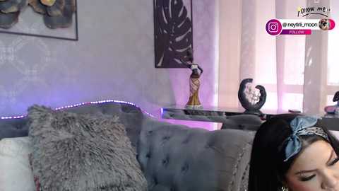 Media: Video of a woman with black hair in a gray tufted couch, wearing a bow headband, in a modern, minimalist bedroom with a white wall, leaf print, and purple LED strip lighting.