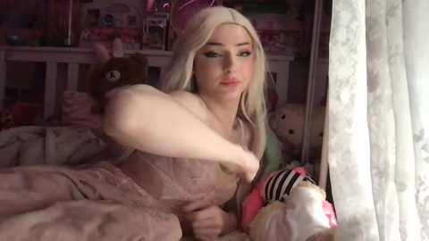 Media: A video of a blonde woman in a light pink dress, hugging a teddy bear, in a cozy bedroom with plush toys and soft, pastel-colored bedding.