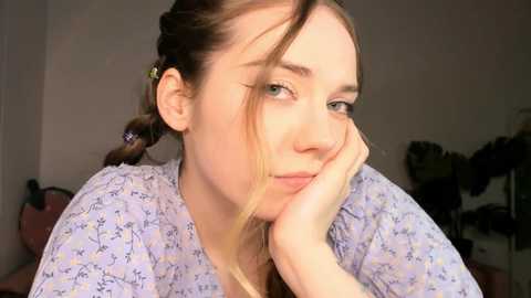 Media: Video of a young Caucasian woman with fair skin, light brown hair in braids, wearing a light purple floral top, resting her chin on her hand, with a contemplative expression.