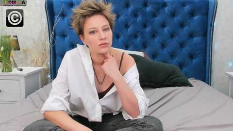Media: Video of a blonde woman in a white shirt and black bra, sitting on a bed with a blue, tufted headboard.