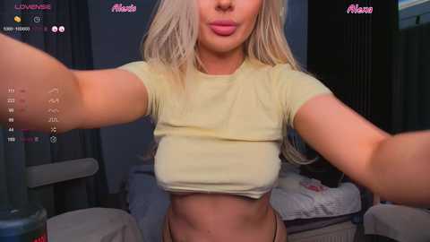 Media: A video of a blonde woman with long hair, wearing a tight yellow crop top, taking a selfie in a dimly-lit room.
