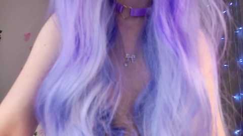 Media: Video of a light-skinned woman with long, wavy hair dyed in shades of lavender and purple. She wears a purple choker and a silver necklace with a small pendant. Background includes a blurred, indoor setting with twinkling lights.