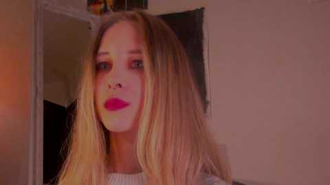 Media: A video of a young, fair-skinned woman with long, straight blonde hair and bright red lipstick. She has a neutral expression, standing indoors with a white wall and a mirror in the background.