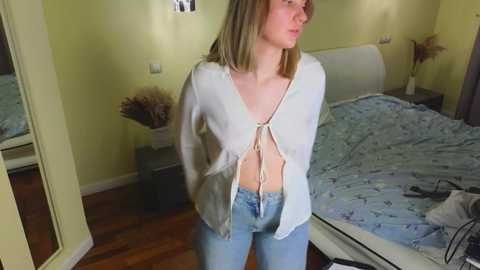 Media: A video of a Caucasian woman with shoulder-length blonde hair, wearing a white, backless, sheer top, and blue jeans, standing in a bedroom with a white bed and yellow walls.