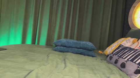 Media: Video of a cozy bedroom with a green bedspread, blue pillows, and a decorative pillow with stripes, set against a dark curtain with a green glow.