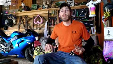 Media: Video of a man in an orange shirt, sitting on a motorcycle in a cluttered garage. Background includes various tools, helmets, and a colorful wall.