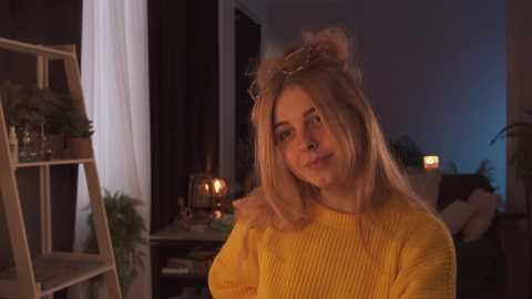 Media: Video of a young woman with blonde hair wearing a yellow sweater, standing in a warmly lit, cozy room with a fireplace, shelves, and a ladder.