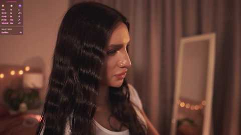 Video of a young woman with long, dark, wavy hair, wearing a white top, standing indoors near a mirror, with soft, warm lighting and a cozy background.