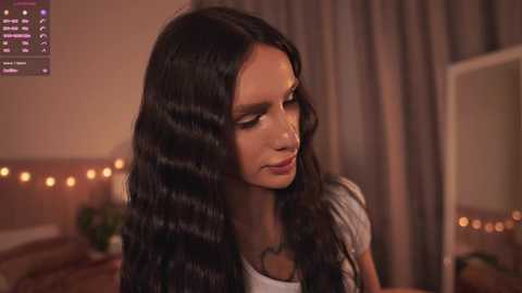 Video of a young woman with long, dark, wavy hair, wearing a white top and necklace, sitting in a dimly lit bedroom with beige curtains and string lights.