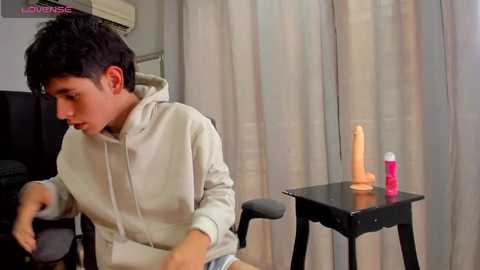 Media: A video of a young boy with short black hair, wearing a beige hoodie, sitting on a black chair. He holds a realistic tan dildo on a black table, near beige curtains.