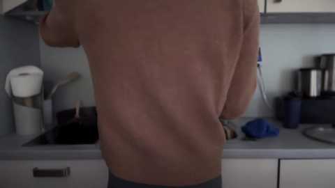 Media: Video of a shirtless man with a tan back, standing in a modern kitchen. He has light skin, and the scene includes a white counter with a coffee maker, kettle, and utensils.
