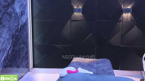 Media: Video of a modern bedroom with a black, geometric-patterned wall, two sconce lights, a grey bed with a white blanket and pink pillow, and a green and white NICHOLAS SHELL watermark.