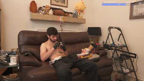 Media: Video of a bearded, shirtless man with a tattoo on his chest, playing a video game on a brown leather couch in a cluttered living room with a Star Wars-themed shelf and a folding chair.