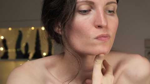 Media: Video of a pale-skinned woman with dark hair in a messy bun, wearing a serene expression, touching her chin. Background features a blurred, decorative golden wall with bohemian-style hanging items.
