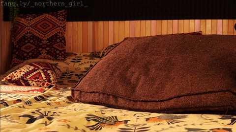 Media: Video of a cozy bed with a textured brown pillow, patterned yellow bedspread, and geometric red and black pillows on a wooden headboard, in a warm, rustic room.