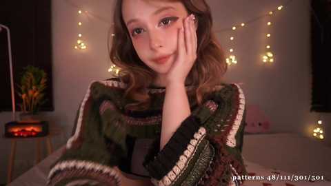 Media: Video of a young woman with fair skin and wavy brown hair, wearing a patterned green sweater, resting her chin on her hand. Warm, ambient lighting with fairy lights and a small plant in the background.