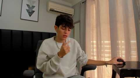 Media: Video of a young man with short dark hair, wearing a white sweatshirt, gesturing with his right hand. He sits in an office chair near a desk with a window and beige curtains.