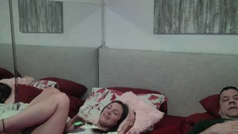 A video depicts a young woman with fair skin, dark hair, and a light floral top lying on a bed with red and pink pillows. A man with short hair and a green shirt is partially visible in the background.