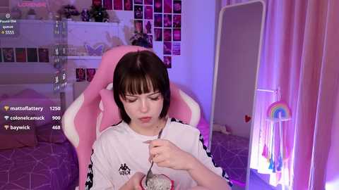 Media: Video of a young woman with pale skin, straight dark hair, wearing a white t-shirt, eating cereal in a pink gaming chair. Background includes a bed, shelves with decorations, and a rainbow lamp.