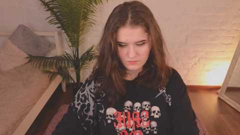 Media: Video of a young woman with long brown hair, wearing a black Metallica t-shirt, seated indoors next to a white brick wall and potted plant.
