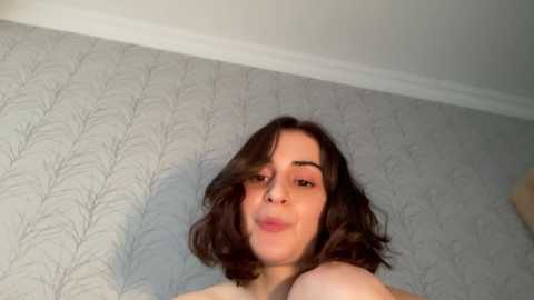 Media: Video of a young woman with medium-length, wavy brown hair, light skin, and a small nose, smiling and puckering her lips. She is nude, and the background features a wallpaper with a subtle, tree-like pattern.
