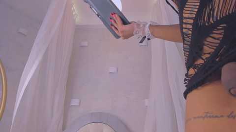 Media: Video of a woman with tan skin, wearing a black lace top and silver bracelet, holding a tablet, standing in a modern bathroom with white tiles and a gold-framed mirror.