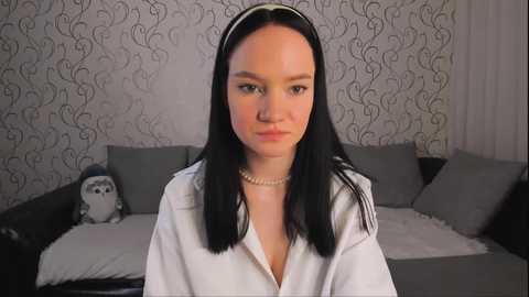 Media: Video of a pale-skinned woman with long black hair, wearing a white robe, in a room with grey and white patterned wallpaper, a grey sofa, and a stuffed bear.